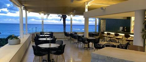 A seaside restaurant with outdoor seating, large windows, and a view of the ocean.