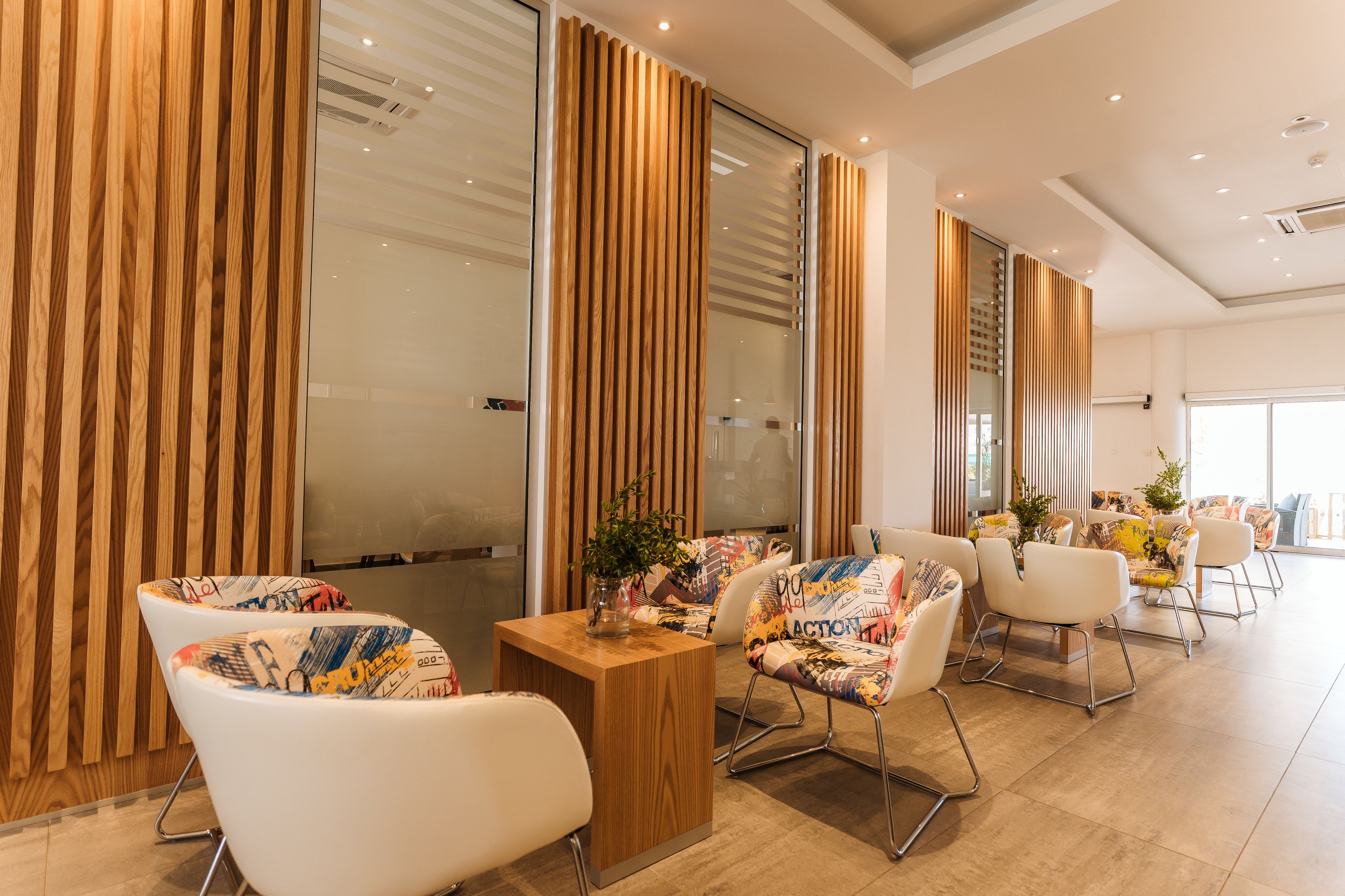 A modern hotel lobby with wooden panel walls, a central wooden table, and colorful patterned chairs.