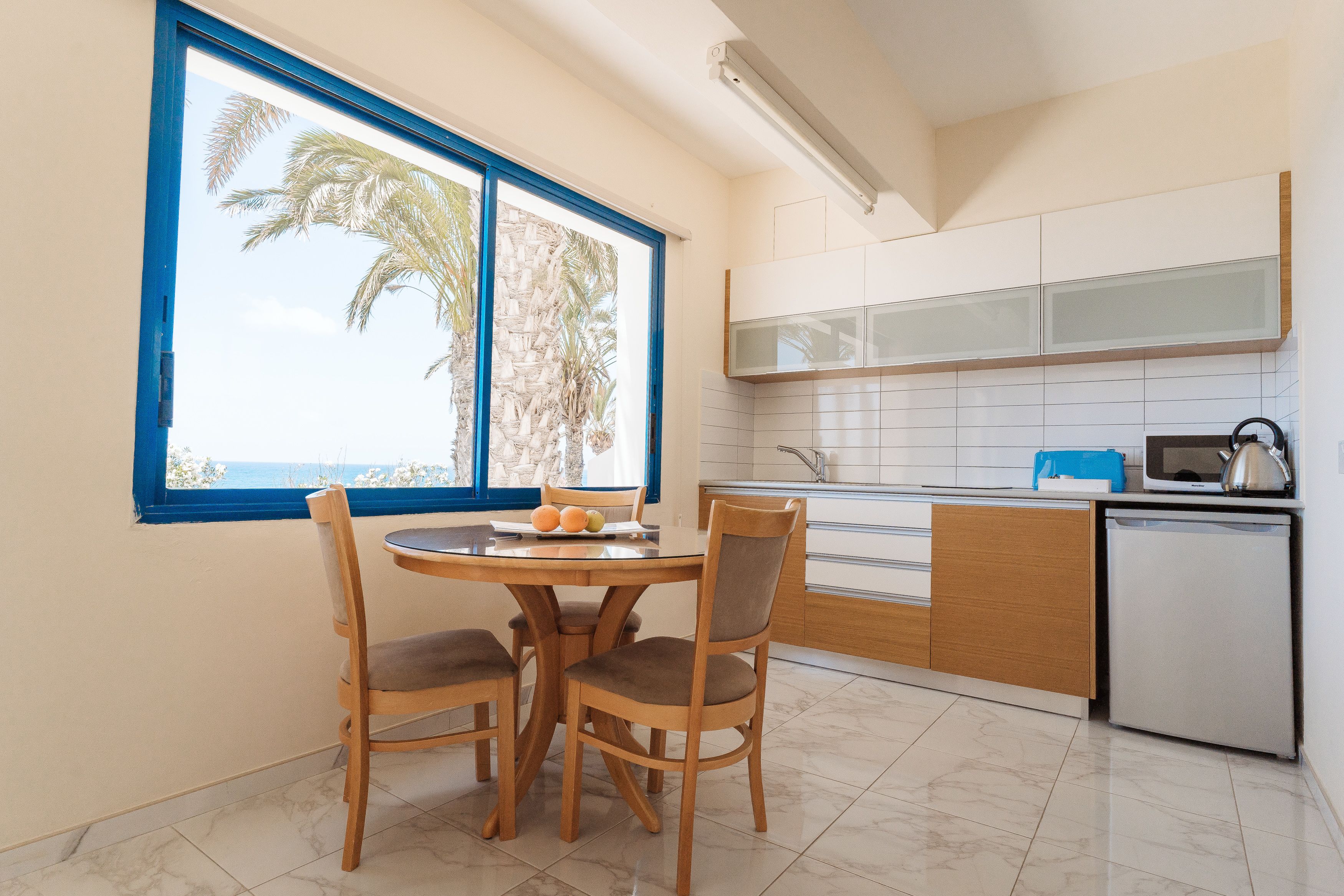 A modern kitchen with a dining area, a window with a view of palm trees, and a small refrigerator.