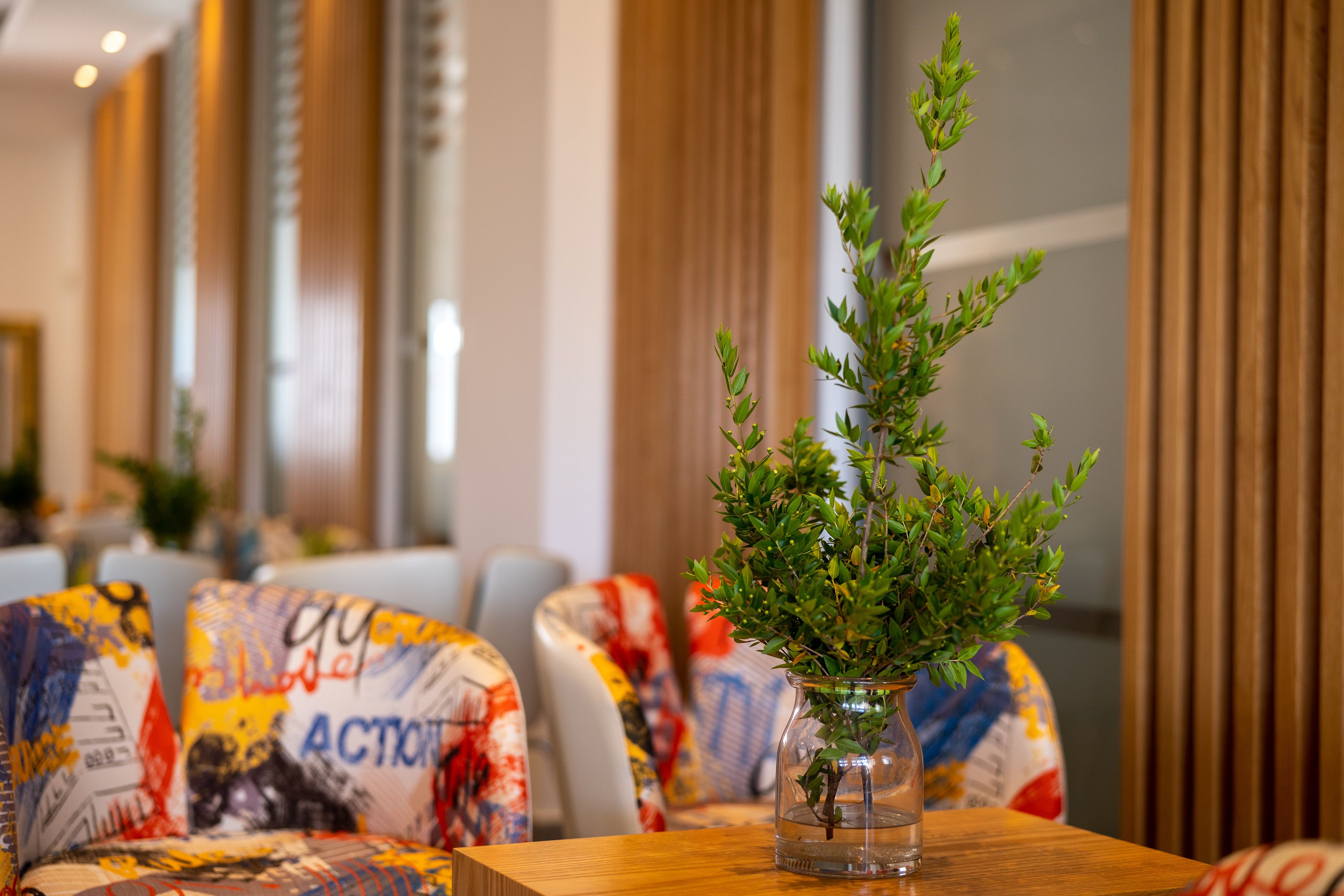 Un interior moderno con una mesa de madera, sillas tapizadas de colores y un jarrón de cristal transparente con follaje verde.
