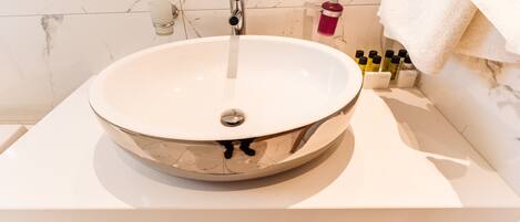 A modern bathroom with a large, round, metallic sink, a marble-tiled wall, and a wooden vanity.