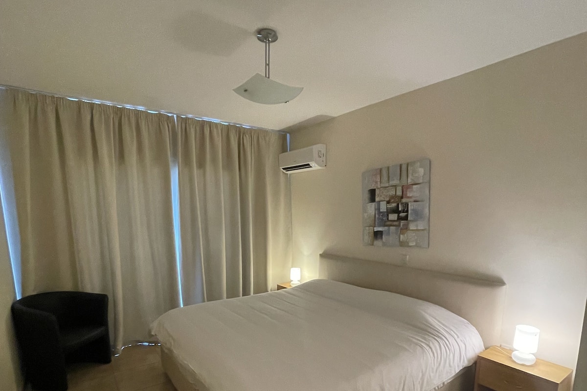 A hotel room with a large bed, a ceiling fan, an air conditioning unit, a bedside table with a lamp, a chair, and a small table.