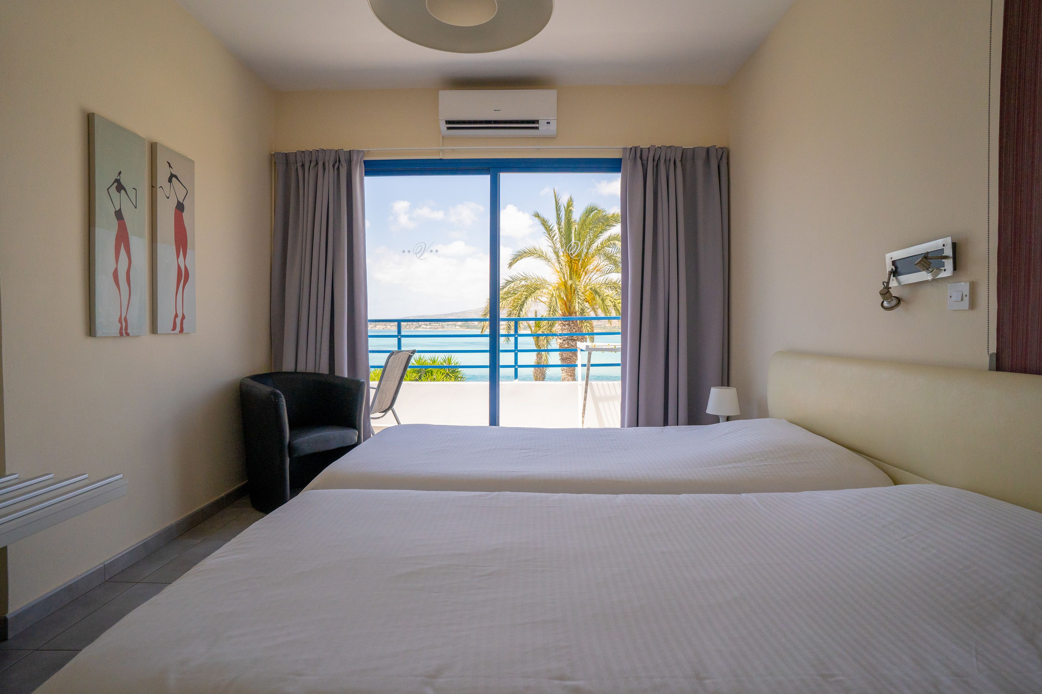 Ein Hotelzimmer mit einem großen Bett, einem Sessel, einem Schreibtisch und Blick auf das Meer und Palmen durch das Fenster.