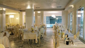 A banquet hall with multiple round tables set for dining, large windows on one side, and a view of the outside. - Aurora (Rimini)