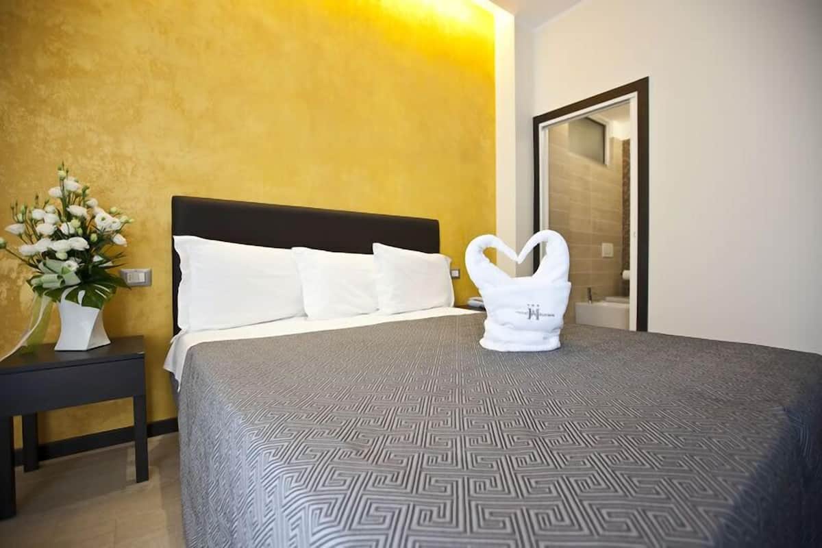 A hotel room with a bed, a nightstand, a vase of flowers, and a towel shaped like a heart on the bed.