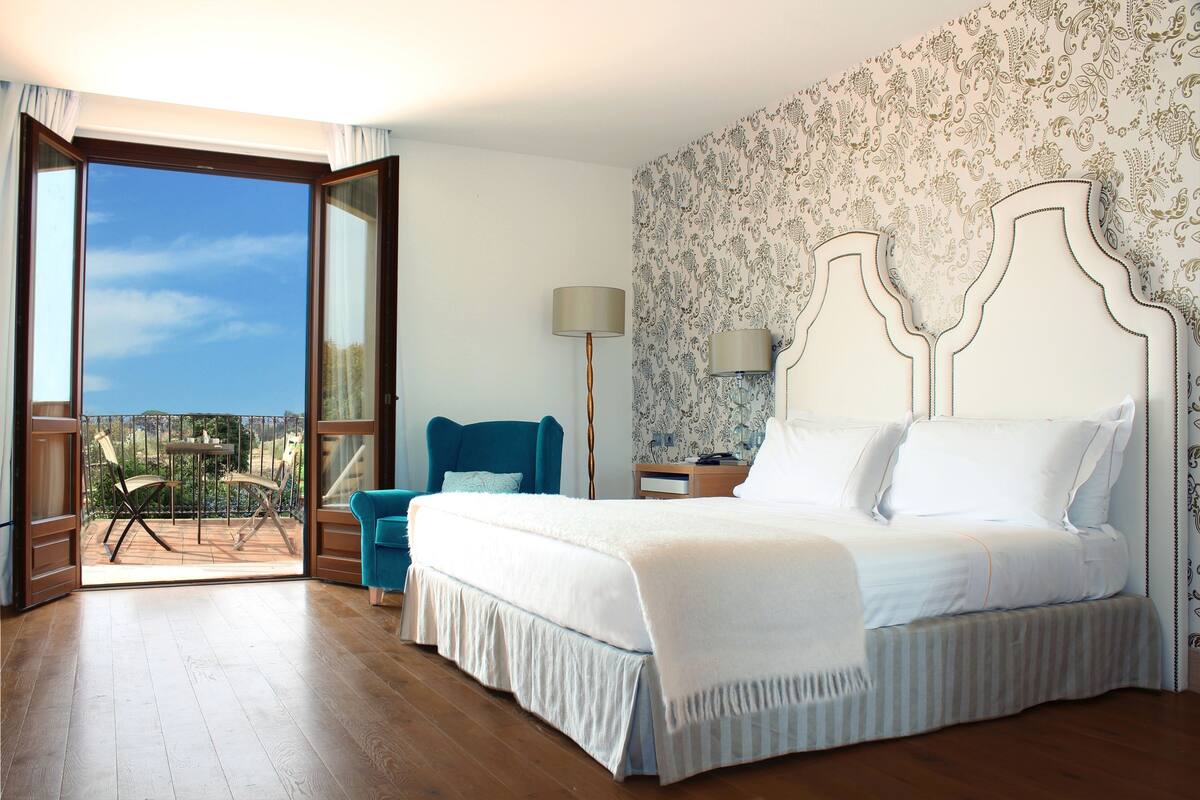 Quarto com uma cama grande, uma cadeira, uma mesinha de cabeceira e vista para o exterior.