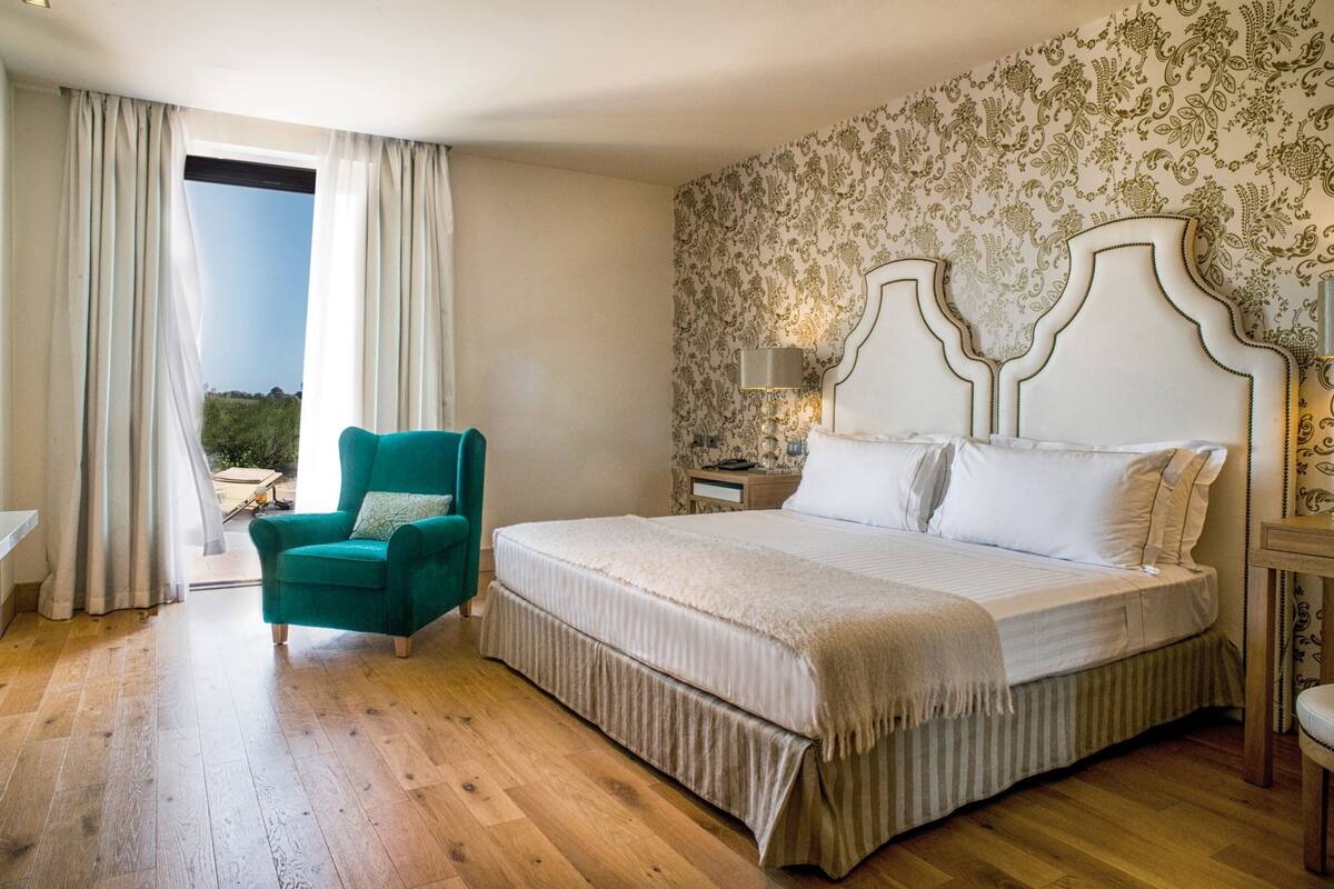 Quarto com cama grande, uma poltrona azul-esverdeada, mesa de cabeceira com abajur e janela com cortinas.