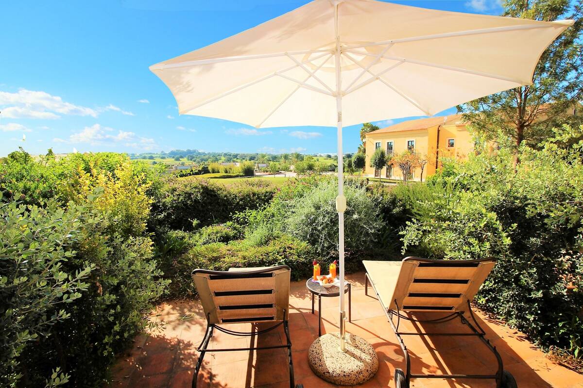 Um pátio externo com duas cadeiras e uma mesa sob um grande guarda-sol, com vista para um jardim ajardinado e um edifício ao fundo.