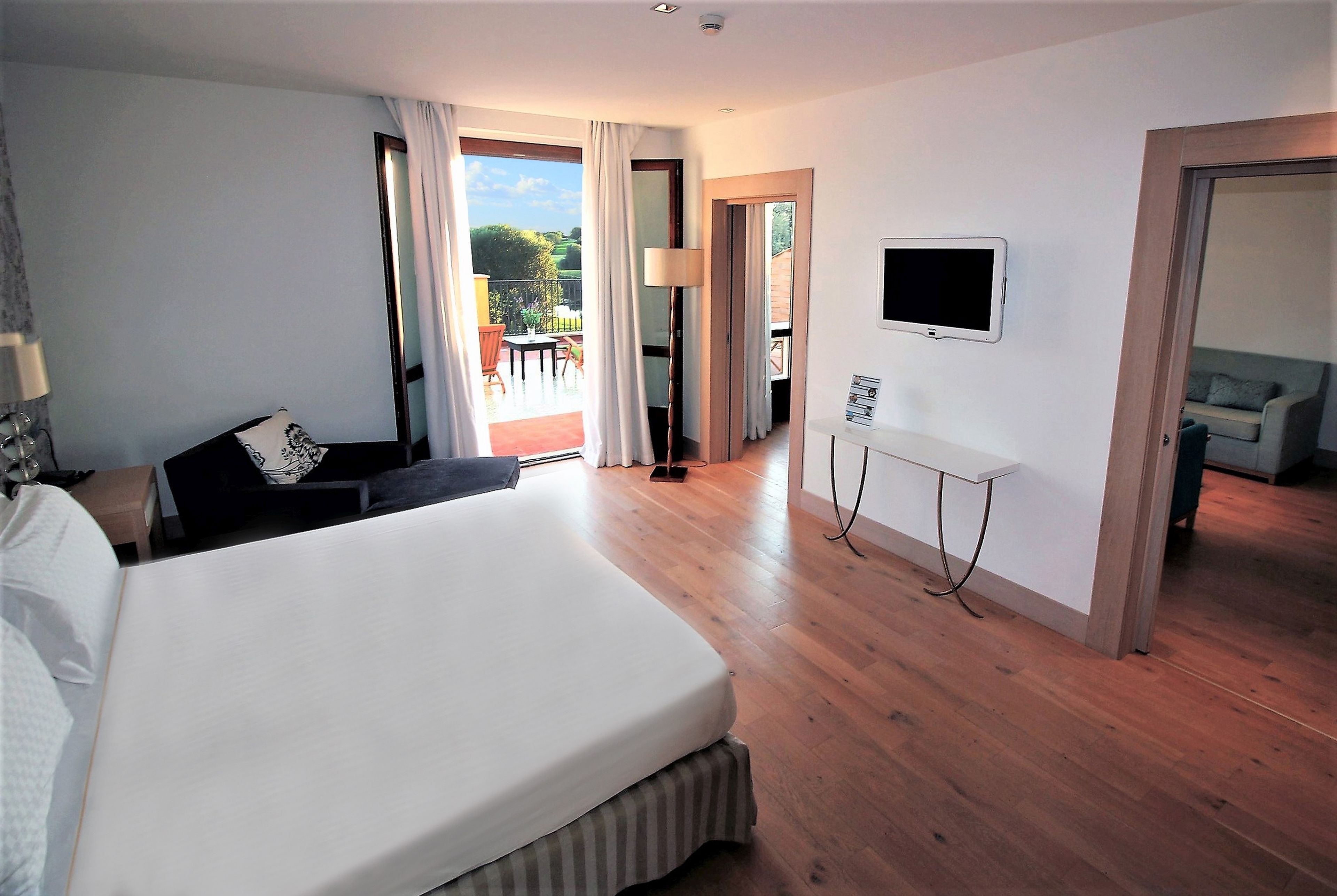 A hotel room with a large bed, a desk, a TV, and a balcony with outdoor furniture.