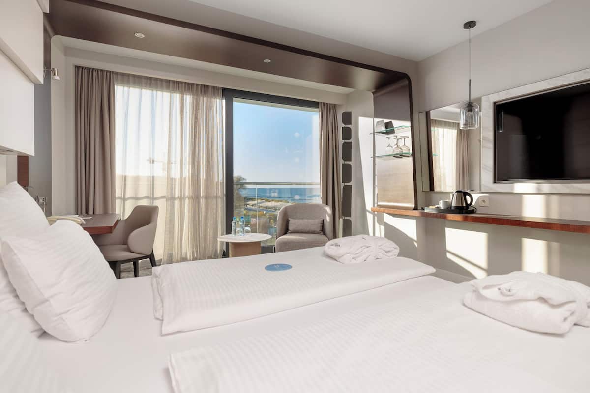 A hotel room with a large bed, a desk, a chair, a small table, a TV, and a view of the city.