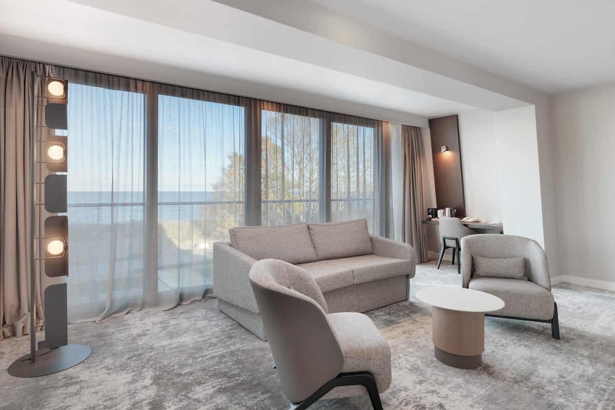 A modern hotel room with a large window, a sofa, two armchairs, a small table, and a desk with a chair.