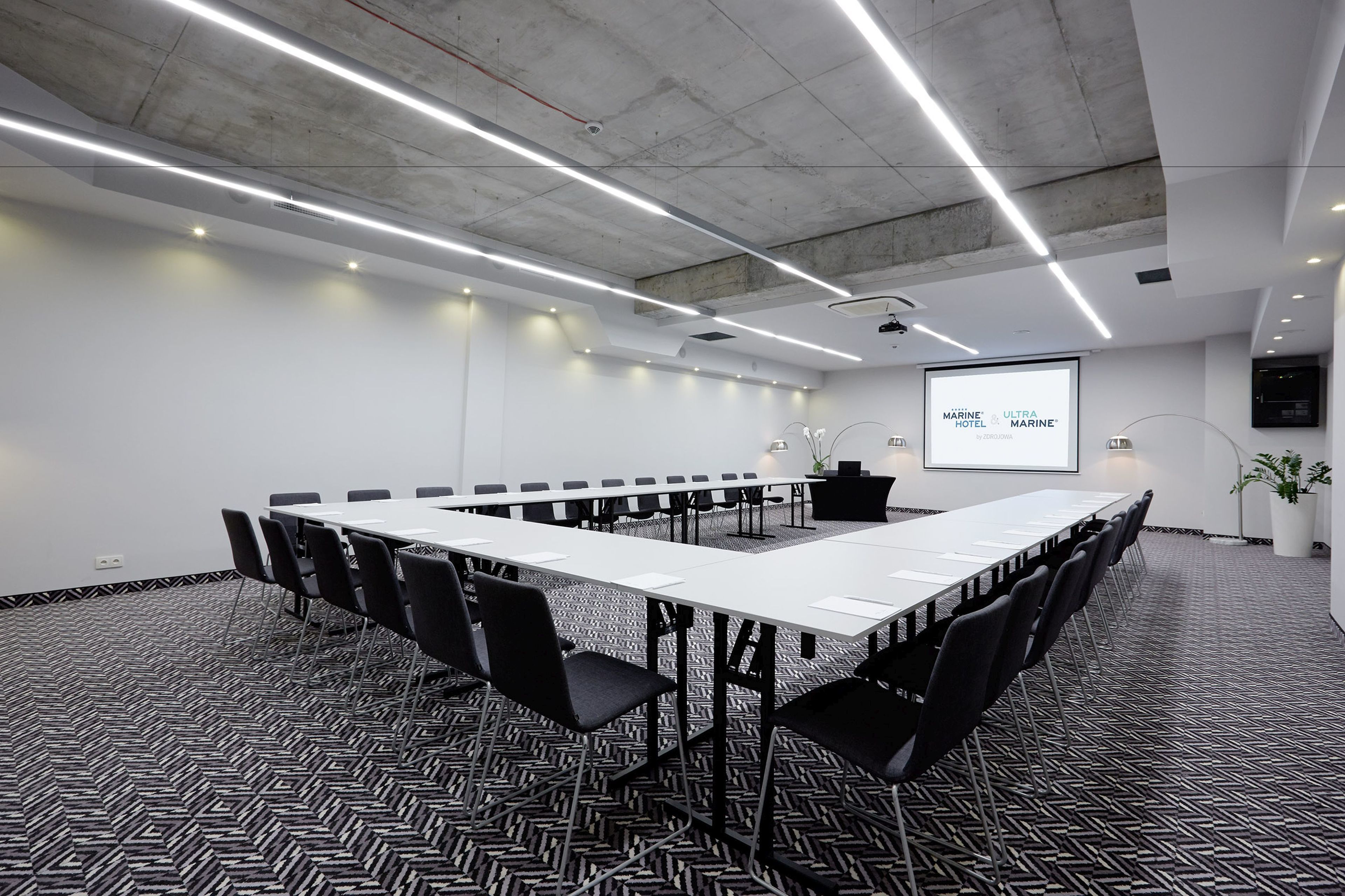 A conference room with a long table, black chairs, a projector screen, and a potted plant.