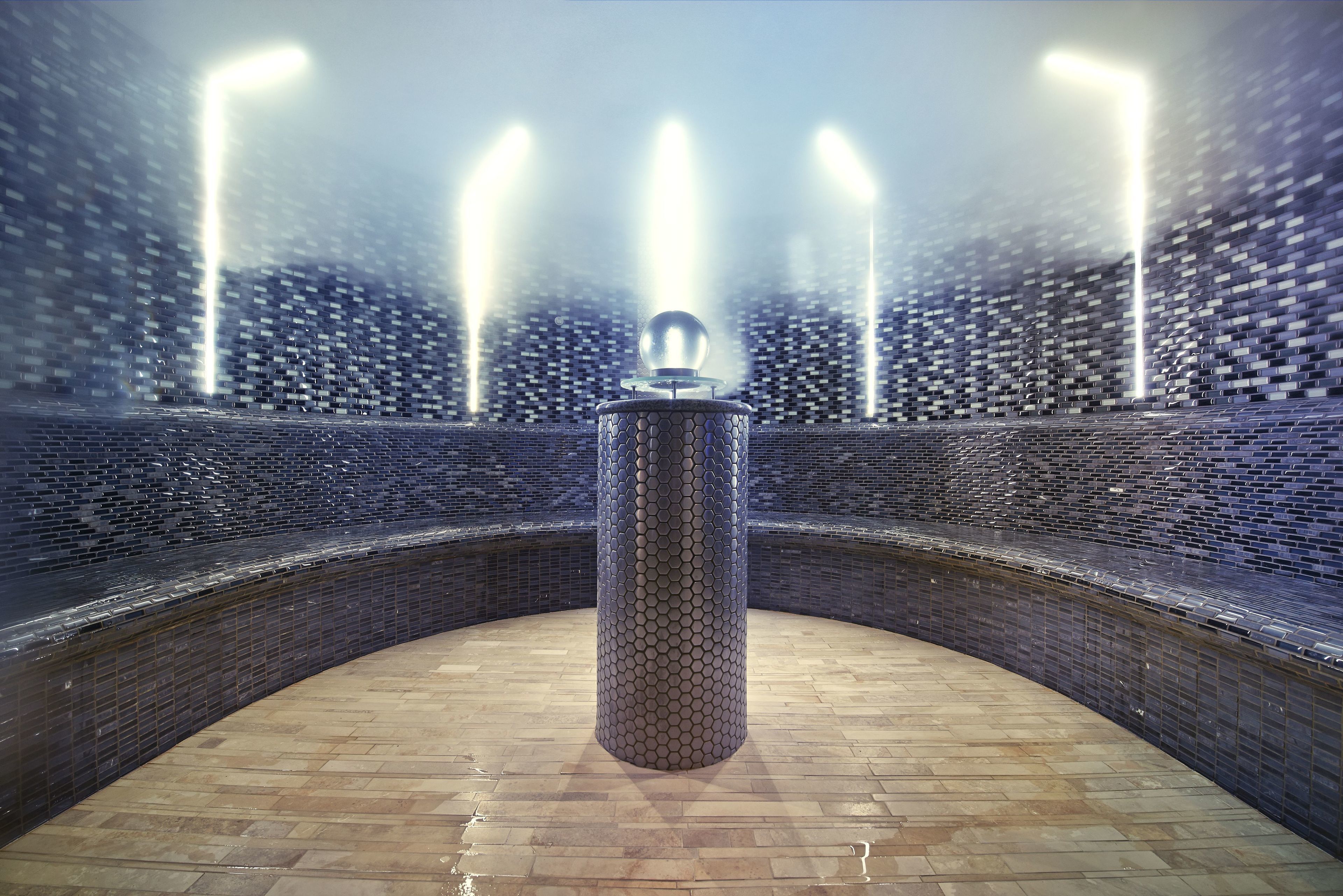 Una fuente circular con una columna central y tres chorros de agua, rodeada por un muro de azulejos.