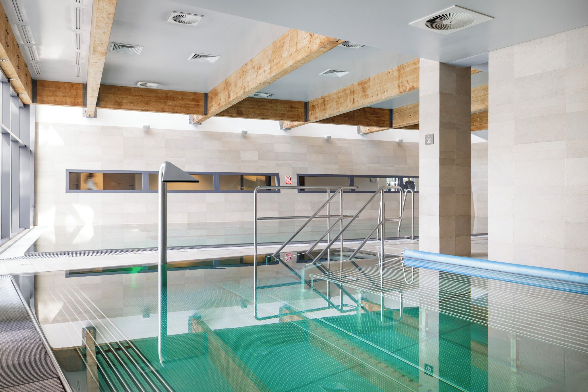 Una piscina interior con paredes de vidrio transparente, vigas de madera en el techo y paredes de azulejos.
