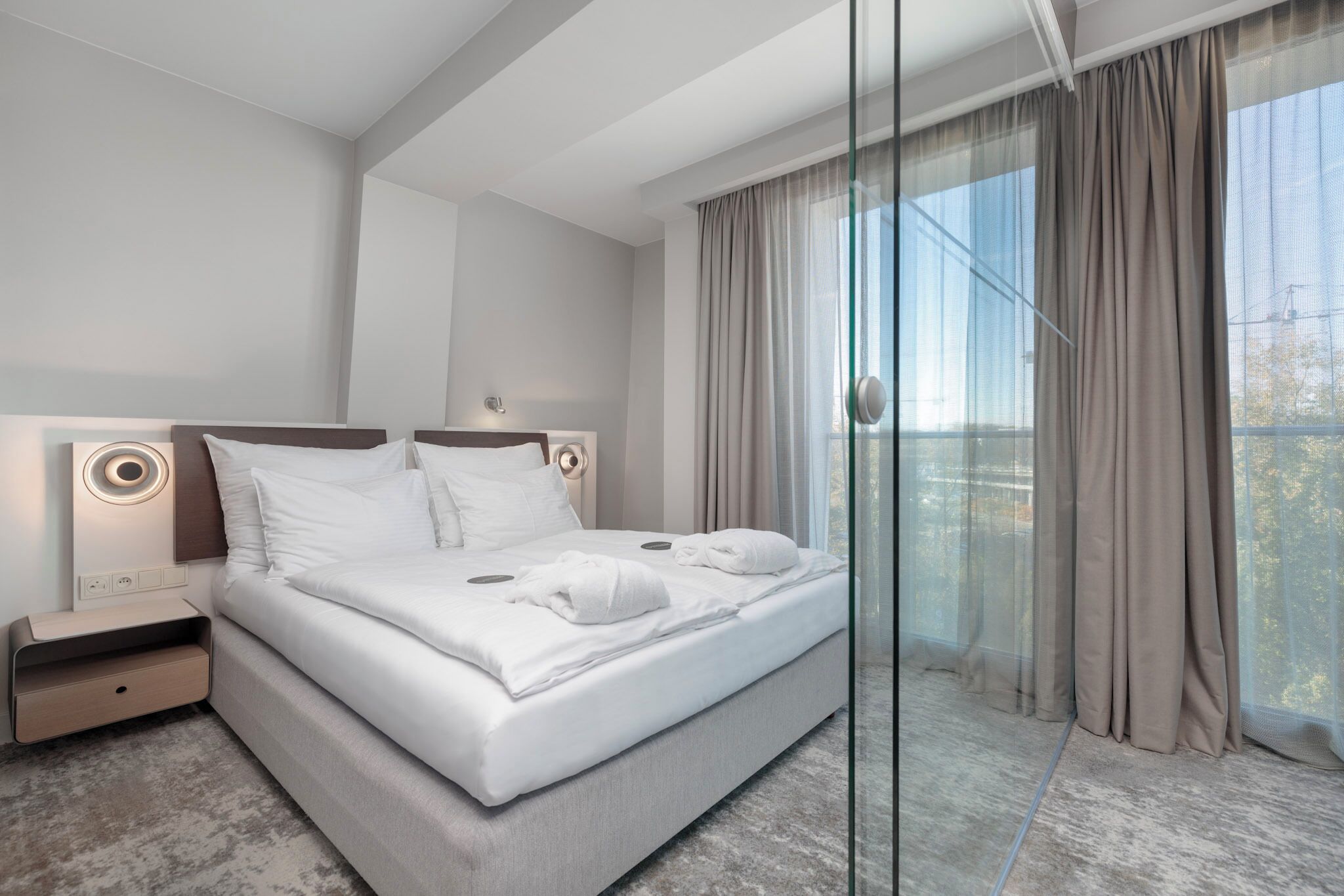 Una camera d'albergo moderna con un letto grande, un comodino e vista attraverso tende leggere.