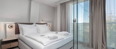 A modern hotel room with a large bed, a bedside table, and a view through sheer curtains.