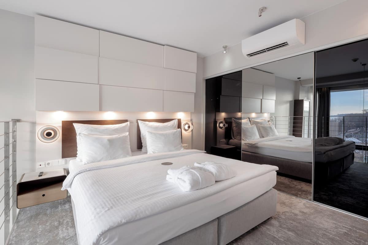A modern hotel room with a large bed, a bedside table, a mirror, and a view of the city.
