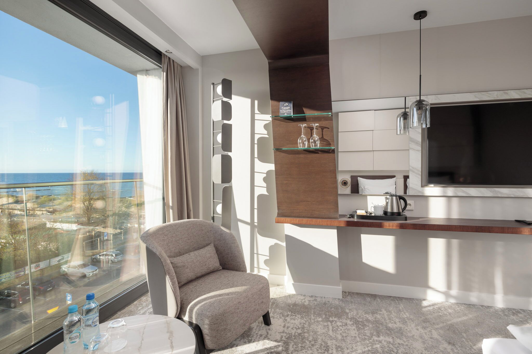 A modern hotel room with a large window, a comfortable armchair, a small table with a water bottle, and a minibar.