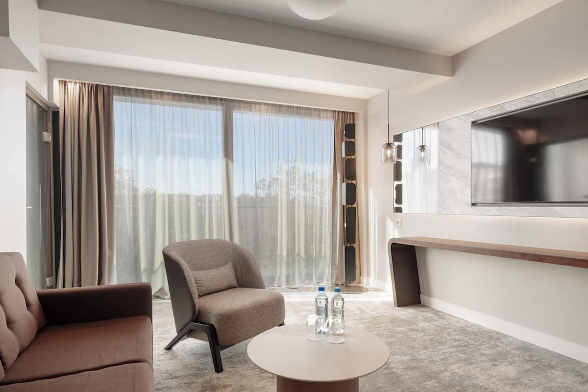 A modern hotel room with a large window, a flat-screen TV, a sofa, a coffee table with water bottles, and a marble accent wall.
