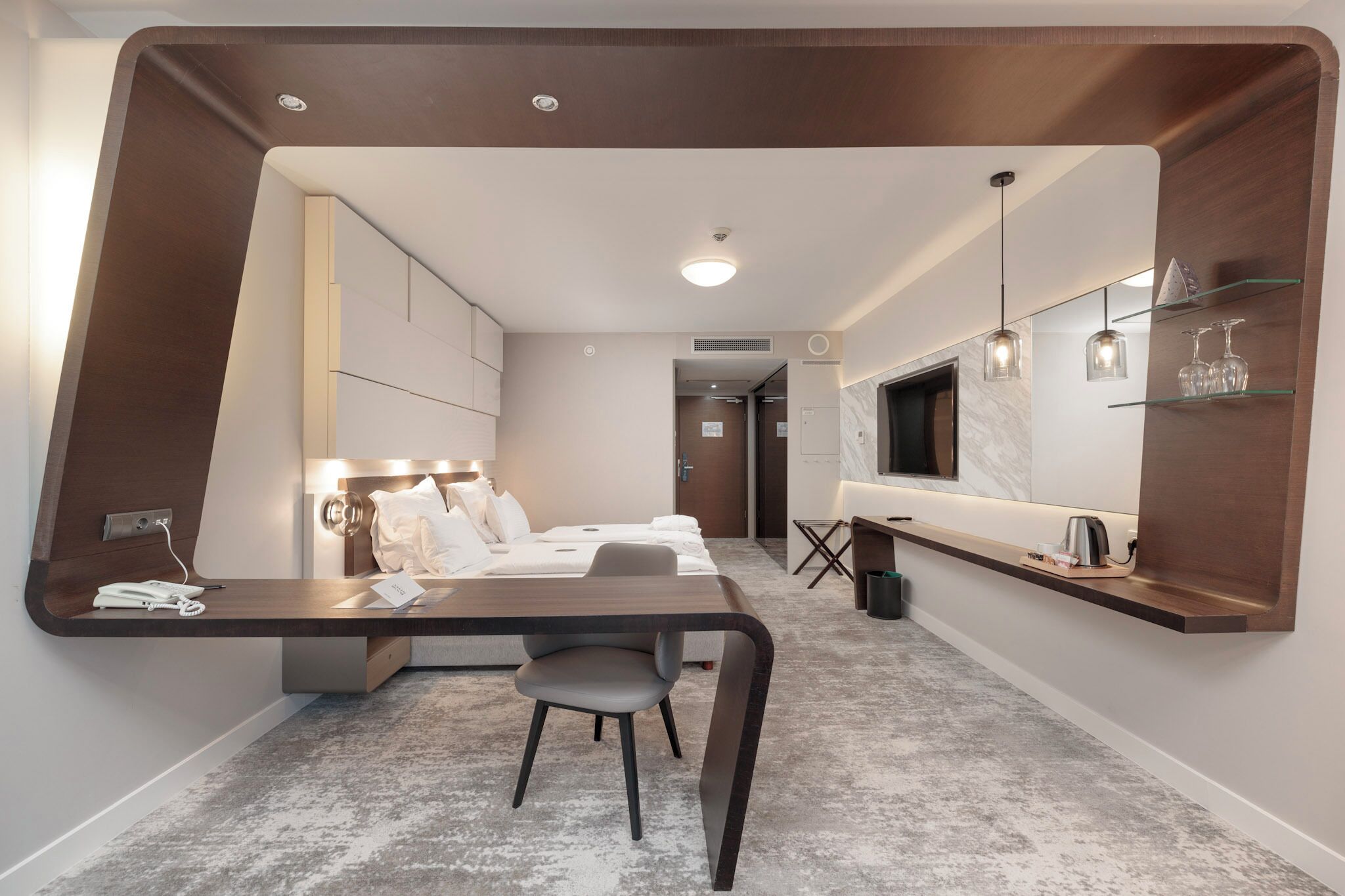 Ein modernes Hotelzimmer mit Bett, Nachttisch, Sessel und Schreibtisch mit Stuhl. An der Wand befindet sich ein großes Spiegel, ein kleiner Tisch mit Wasserkocher und ein Regal mit Gläsern.