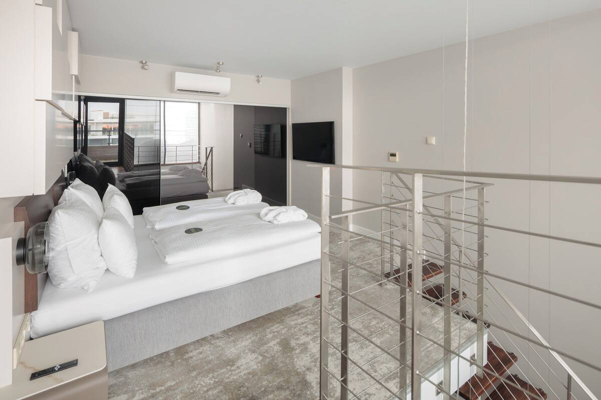 A modern hotel room with a large bed, a glass partition, a flat-screen TV, and a balcony with a view.