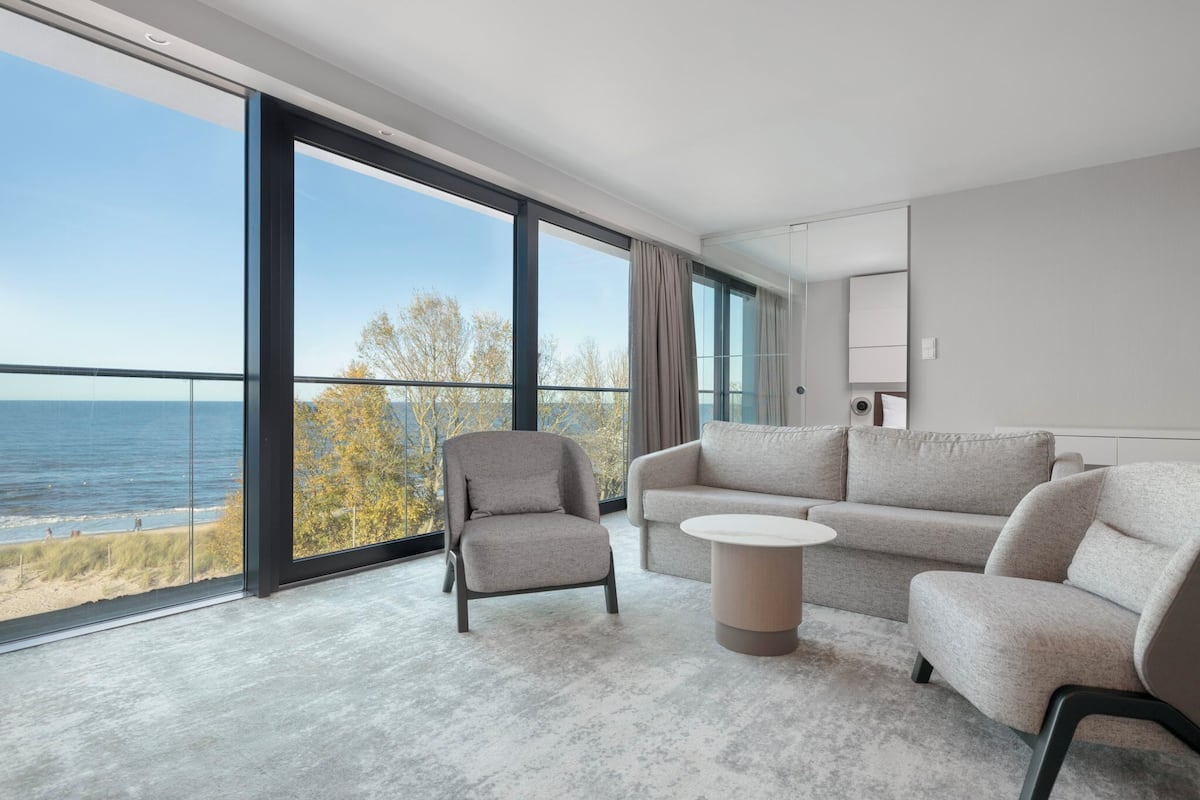 A modern living room with a large window, a grey sofa, a grey armchair, and a small round table.