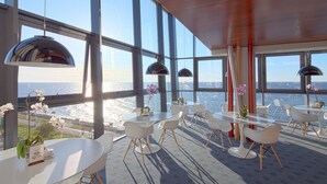Un comedor moderno con grandes ventanales con vista al mar, mesas y sillas blancas y orquídeas decorativas.
