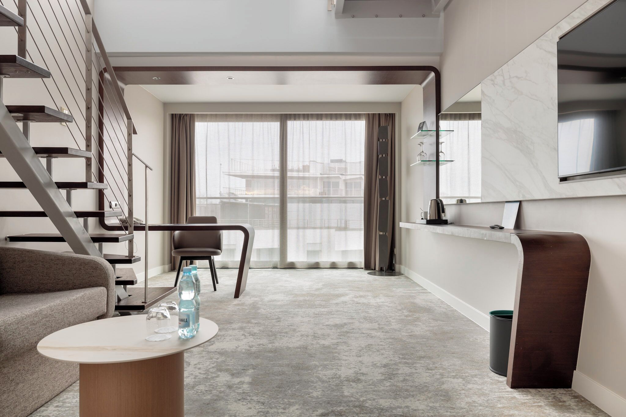 Un salon moderne avec un escalier, un canapé, une table basse et une grande fenêtre avec des rideaux.