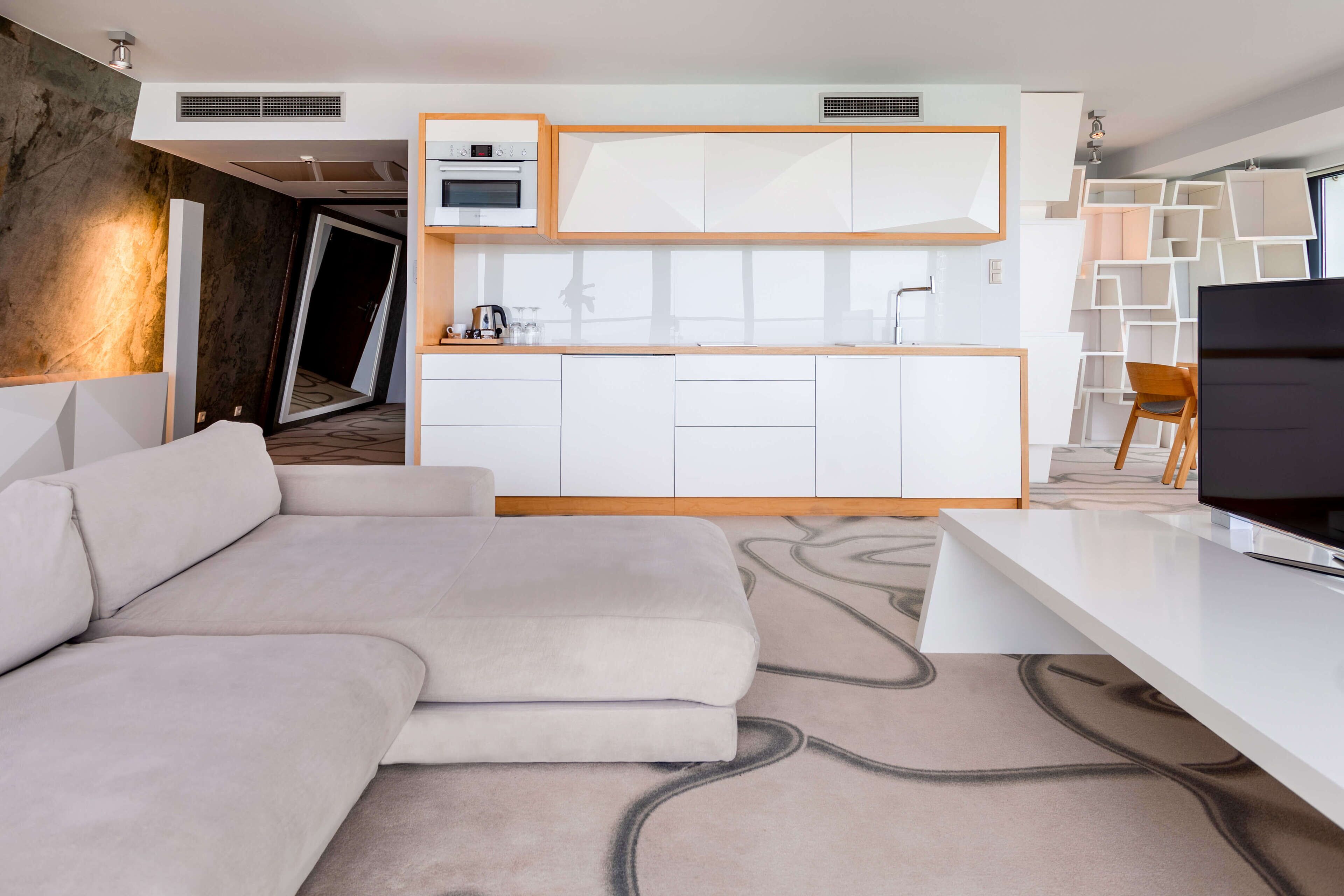 A modern living room with a sofa, a flat-screen TV, and a kitchen area with white cabinets and a built-in oven.