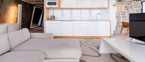 A modern living room with a sofa, a flat-screen TV, and a kitchen area with white cabinets and a built-in oven.