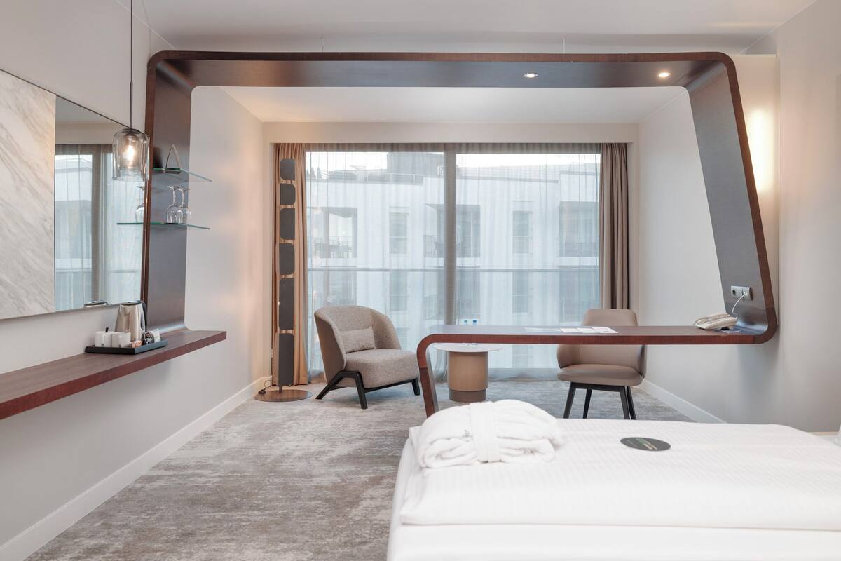 A modern hotel room with a large bed, a desk, a chair, a large window, and a mirror.