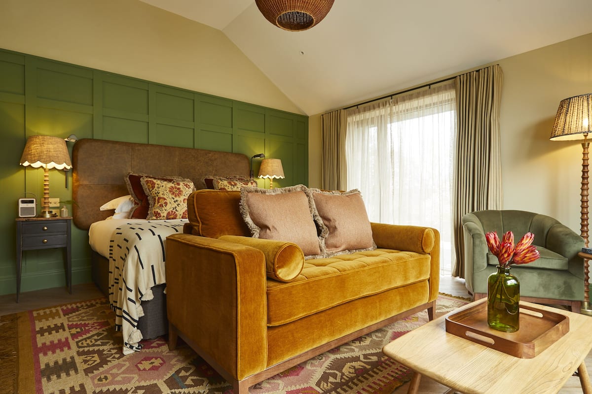 A room with a bed, a sofa, a chair, a wooden coffee table with a vase, and a patterned rug.