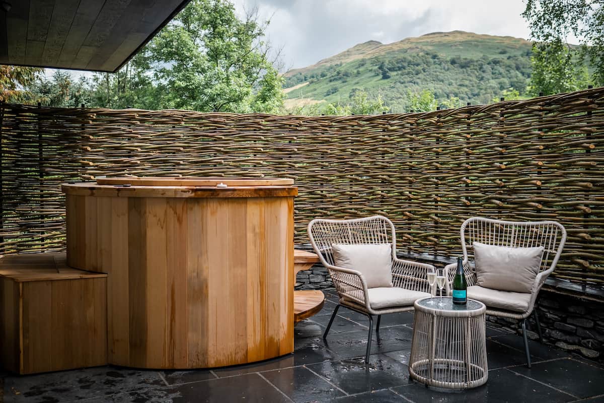 A wooden bar area with wicker chairs and a round table, set against a backdrop of a woven wall and a mountainous landscape.