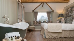 A bedroom with a large bathtub, a bed with multiple pillows, a small stool with toiletries, and a window with curtains. - Rothay Manor Hotel (Ambleside)