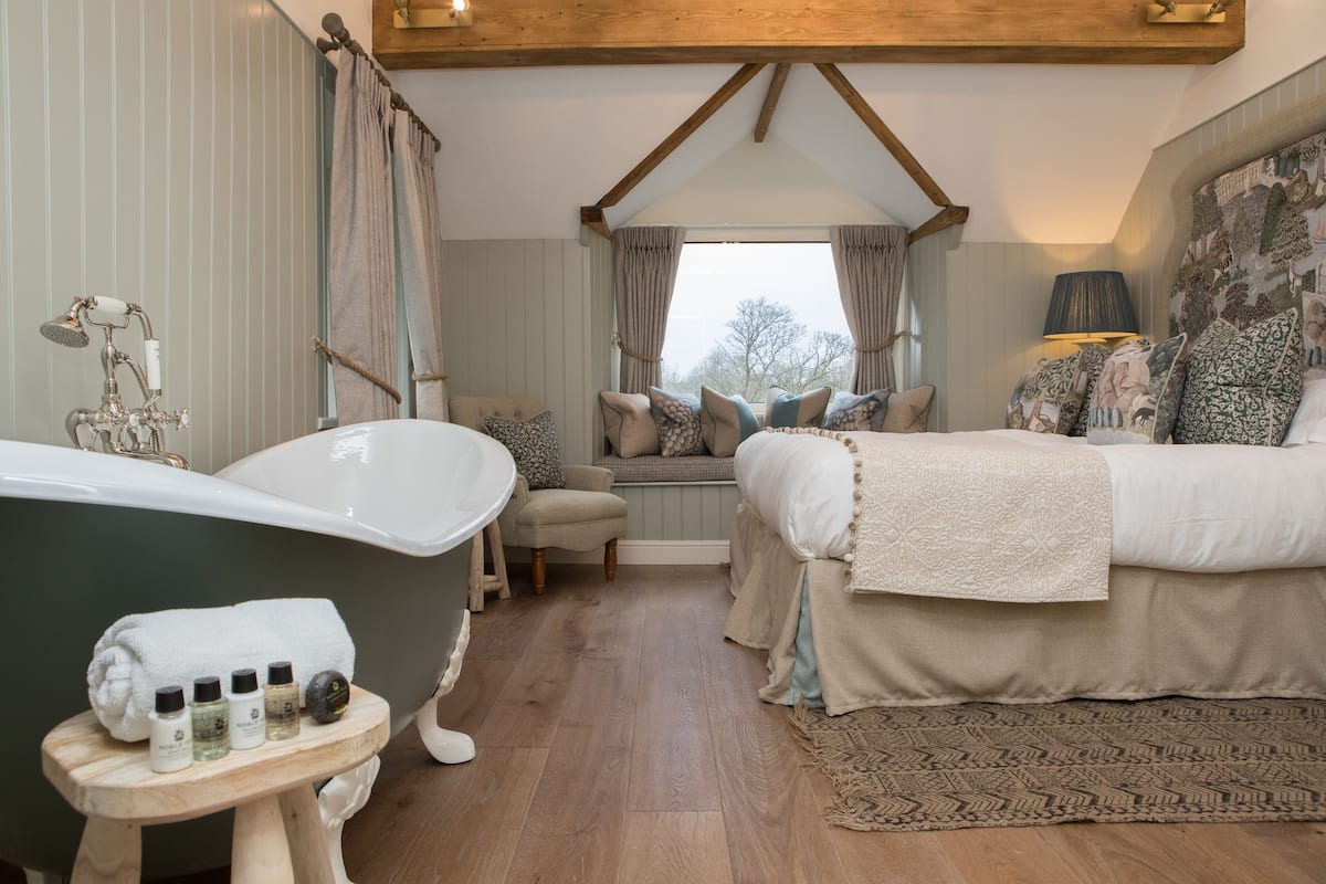 A bedroom with a large bathtub, a bed with multiple pillows, a small stool with toiletries, and a window with curtains.