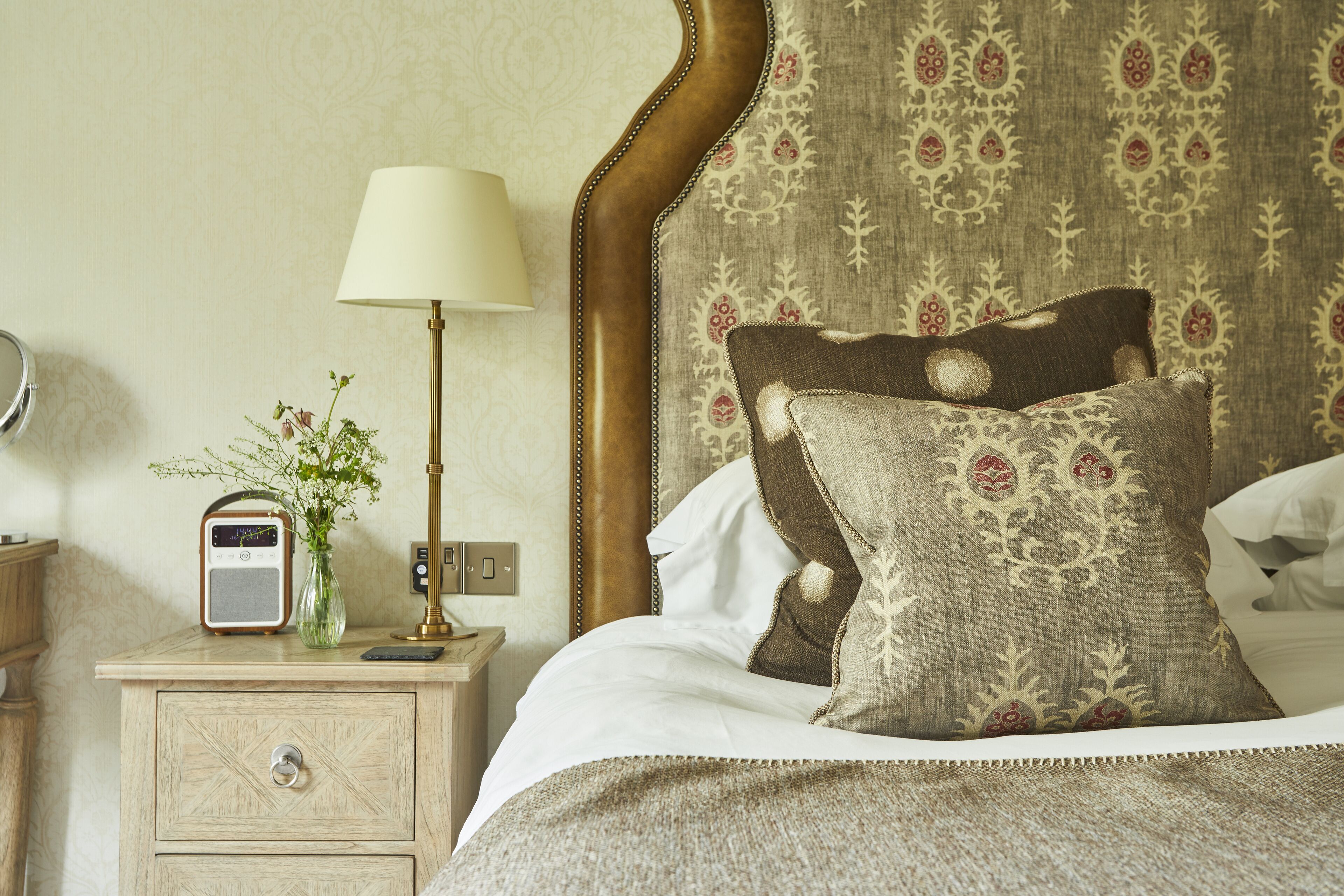 Una cama bien hecha con un cabecero estampado, una mesita de noche con lámpara, radio y un florero con flores.
