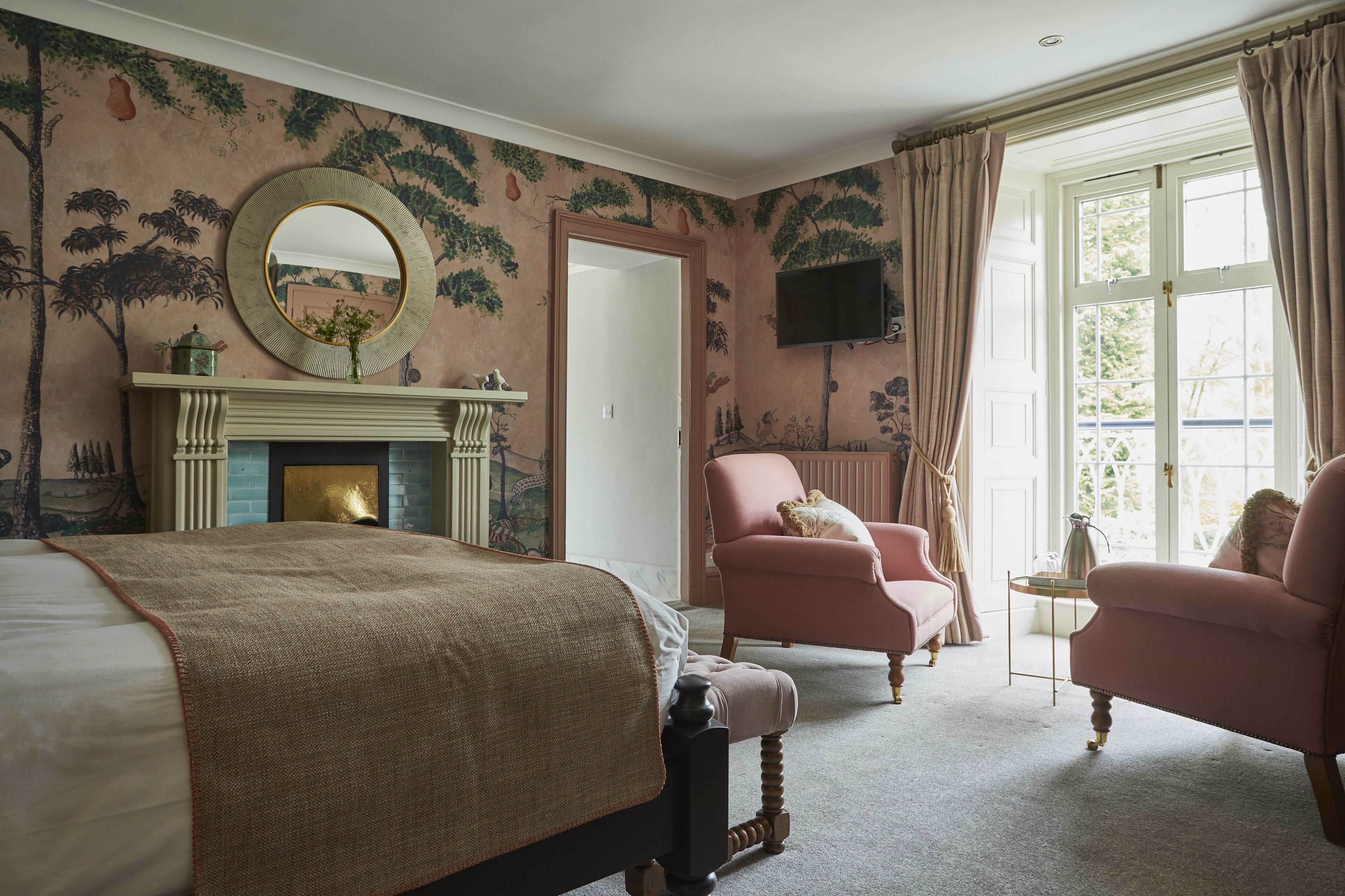 A bedroom with a large bed, a fireplace, two armchairs, a television, and a window with curtains.
