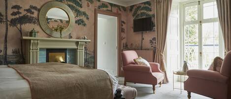 A bedroom with a large bed, a fireplace, two armchairs, a television, and a window with curtains.