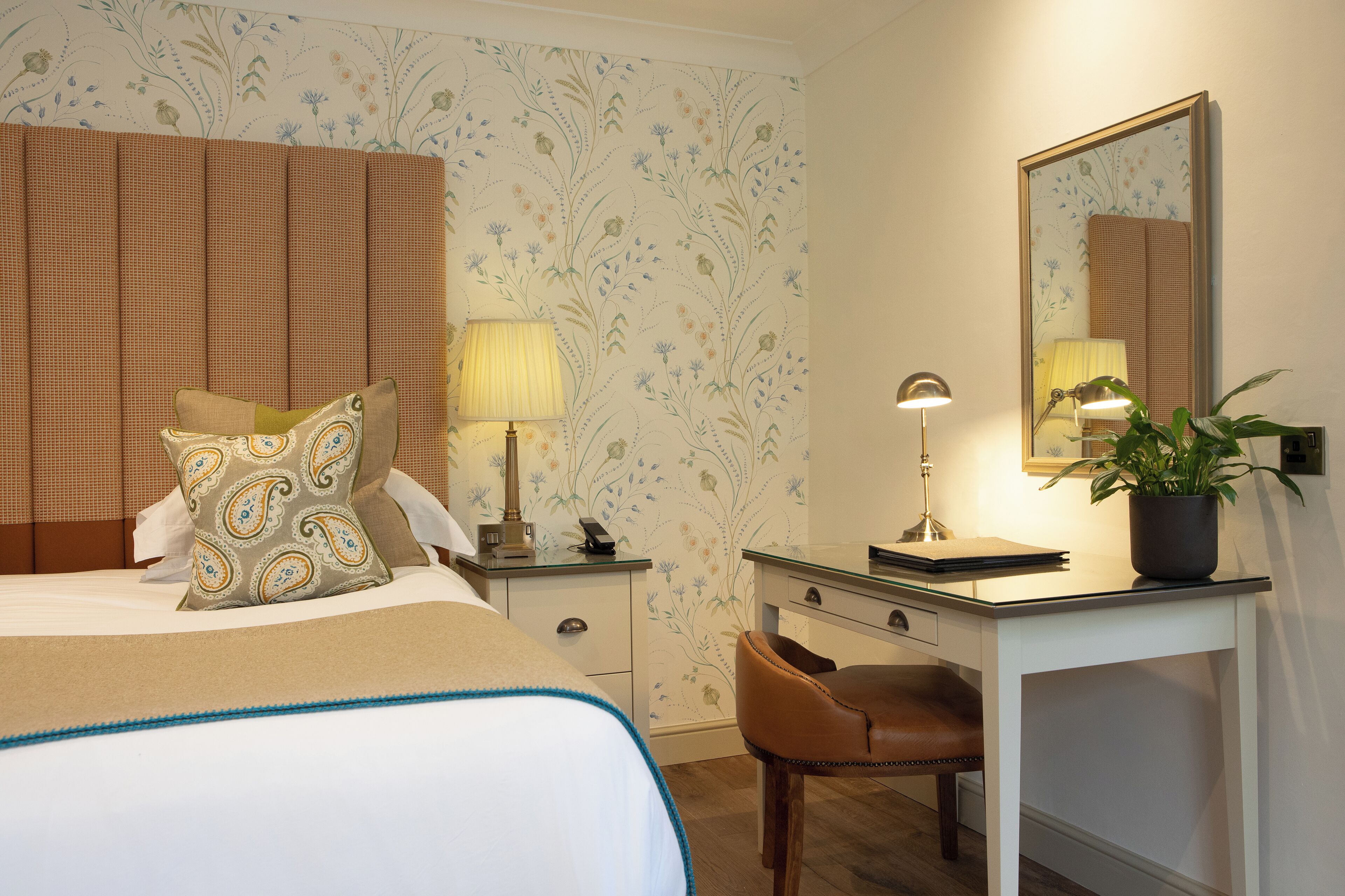 A hotel room with a bed, a desk, a chair, a lamp, a mirror, and a plant.