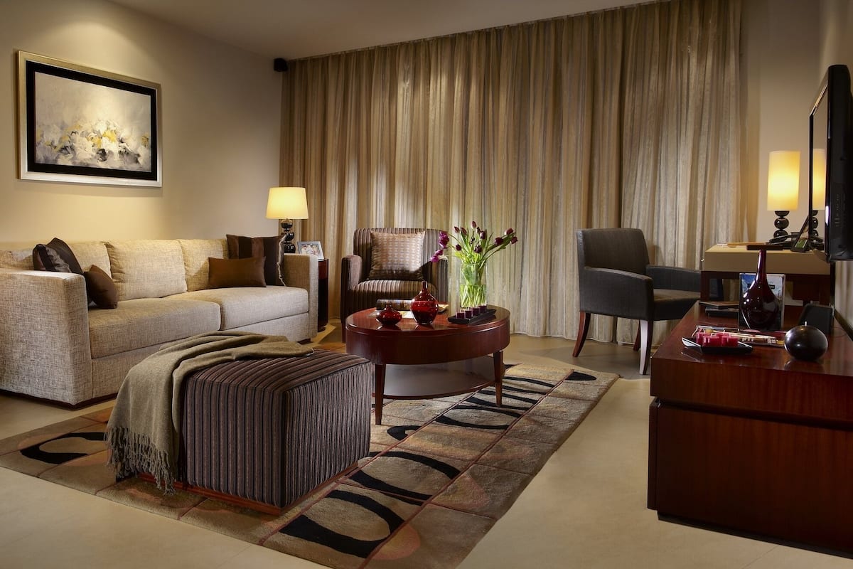 A well-furnished living room with a sofa, armchair, ottoman, and a coffee table. There is a desk with a lamp and a vase of flowers. The room has a large window with curtains.