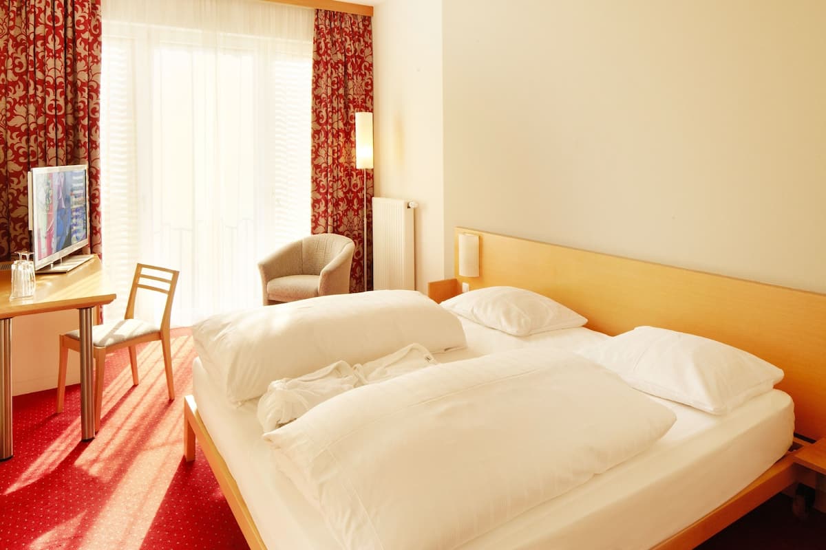 Quarto de hotel com cama, escrivaninha, cadeira, televisão e janela com cortinas.