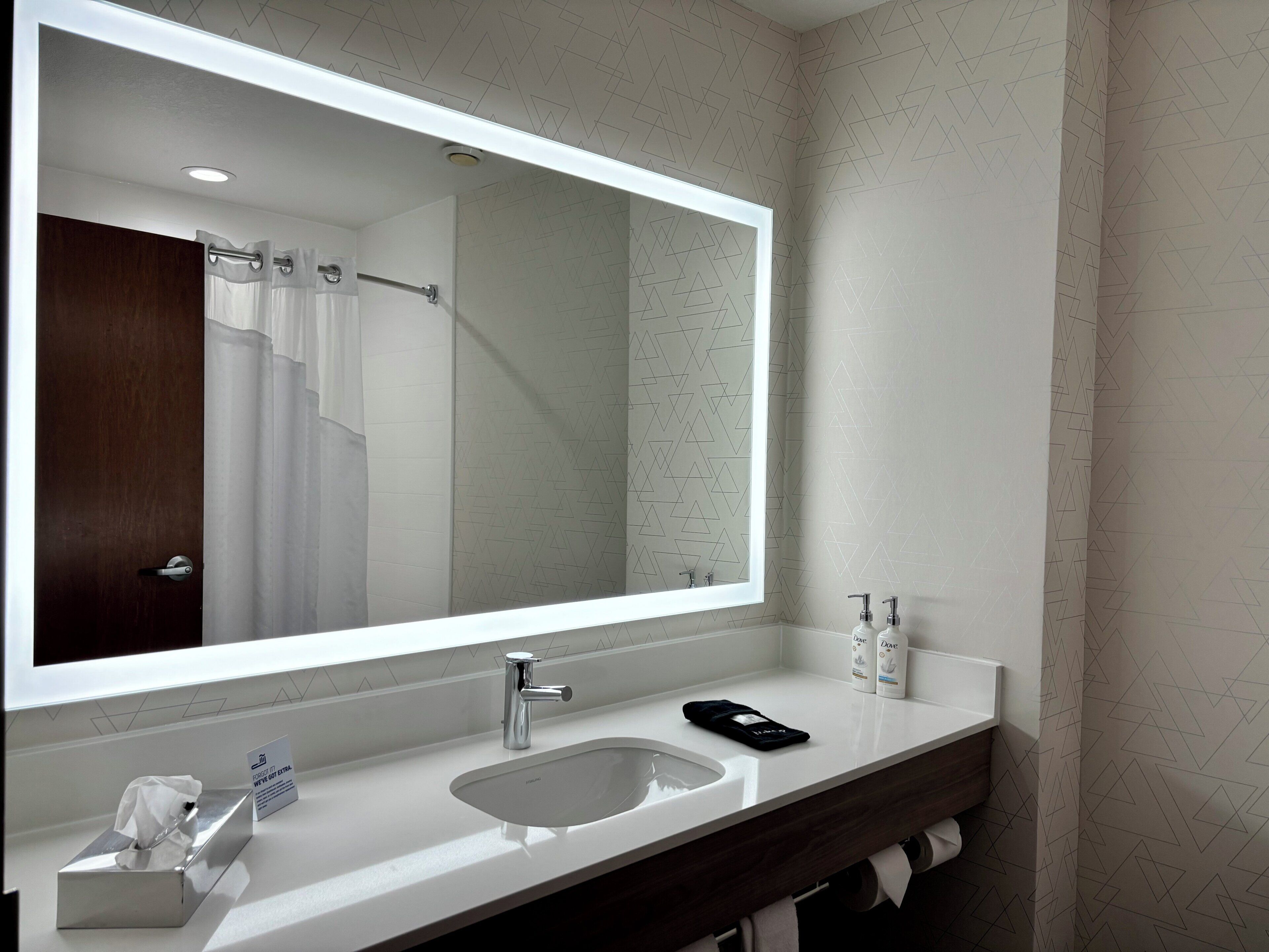 Un baño con un espejo grande, un lavabo, un perchero para toallas y una cortina de ducha.