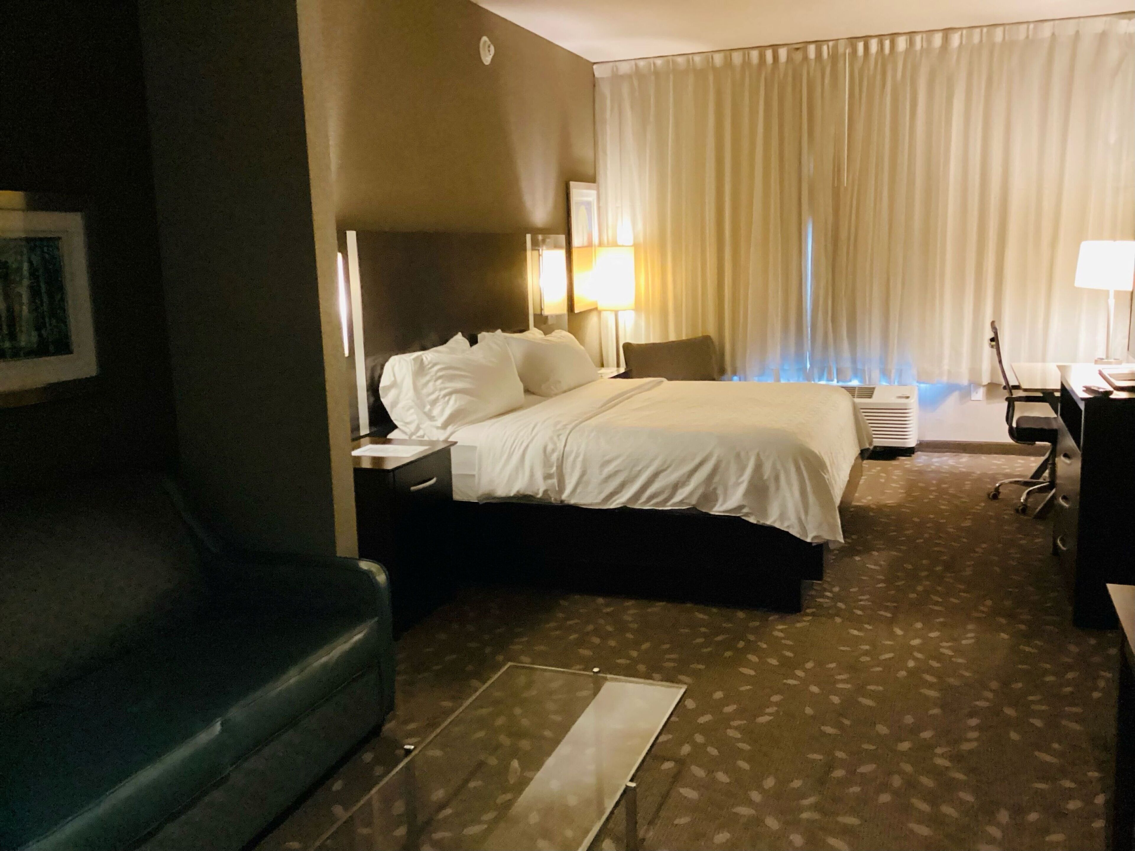 Una habitación de hotel con una cama, un escritorio, una silla, un sofá y una ventana con cortinas.