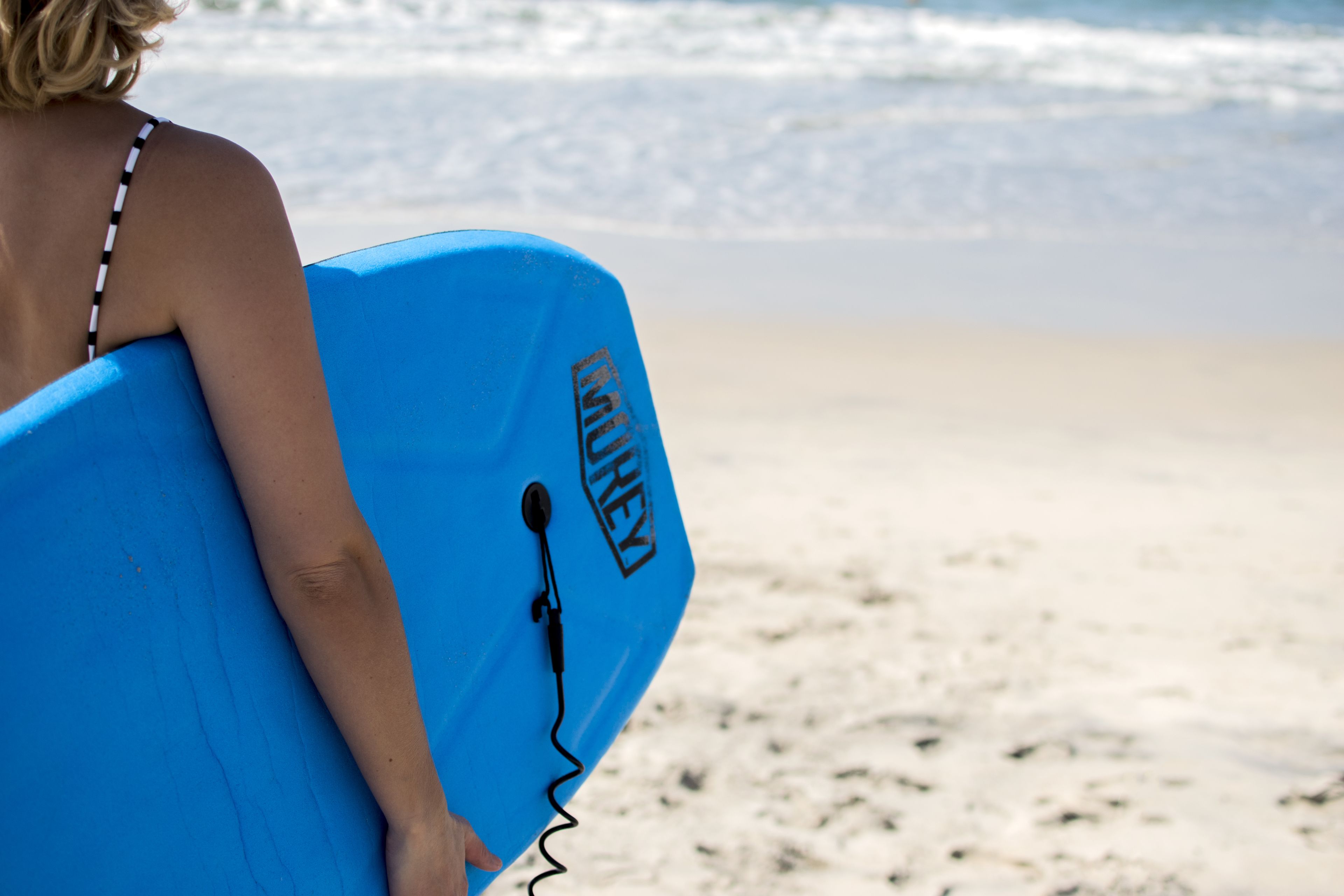 Una persona lleva una tabla de surf azul con un nombre de marca, caminando sobre una playa arenosa.