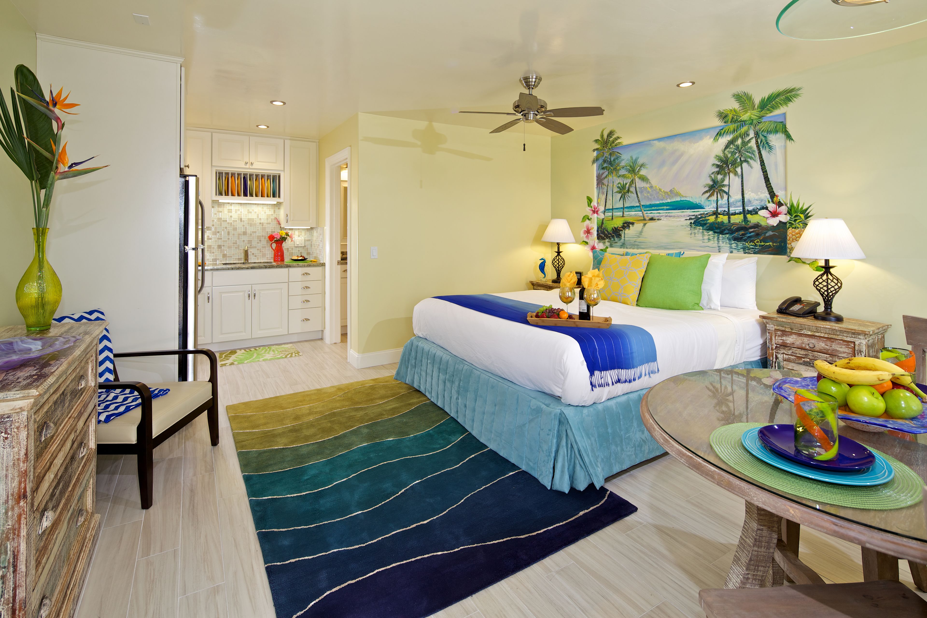 A hotel room with a bed, a dining table, a chair, a kitchen area, and a painting of a tropical scene.