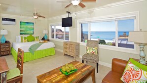 A hotel room with a bed, a desk, a chair, a table, a ceiling fan, a TV, and a view of the ocean. - Ocean Palms Beach Resort (Carlsbad)