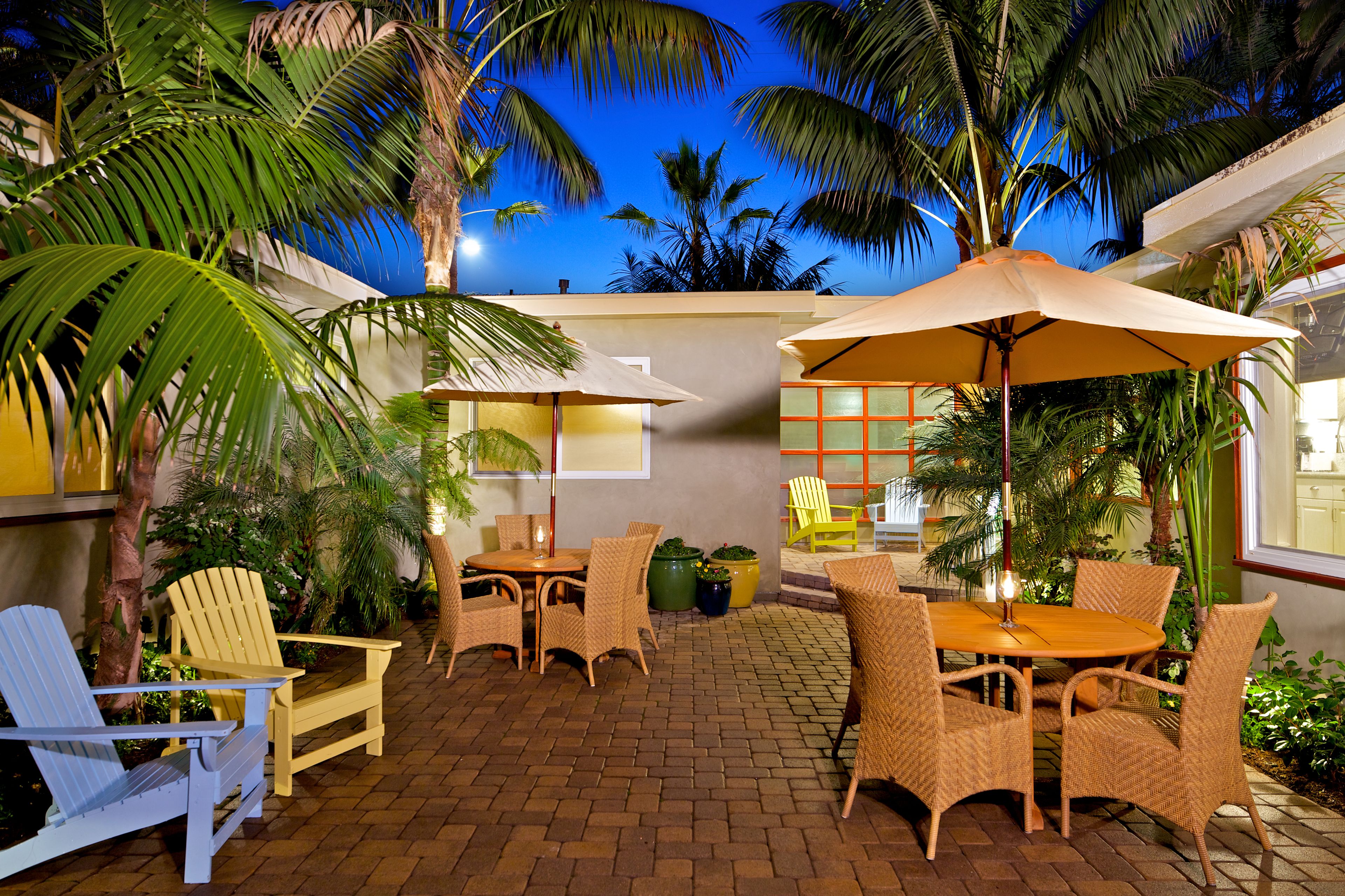 An outdoor patio with wicker furniture, a table, and chairs, surrounded by palm trees and a brick floor.