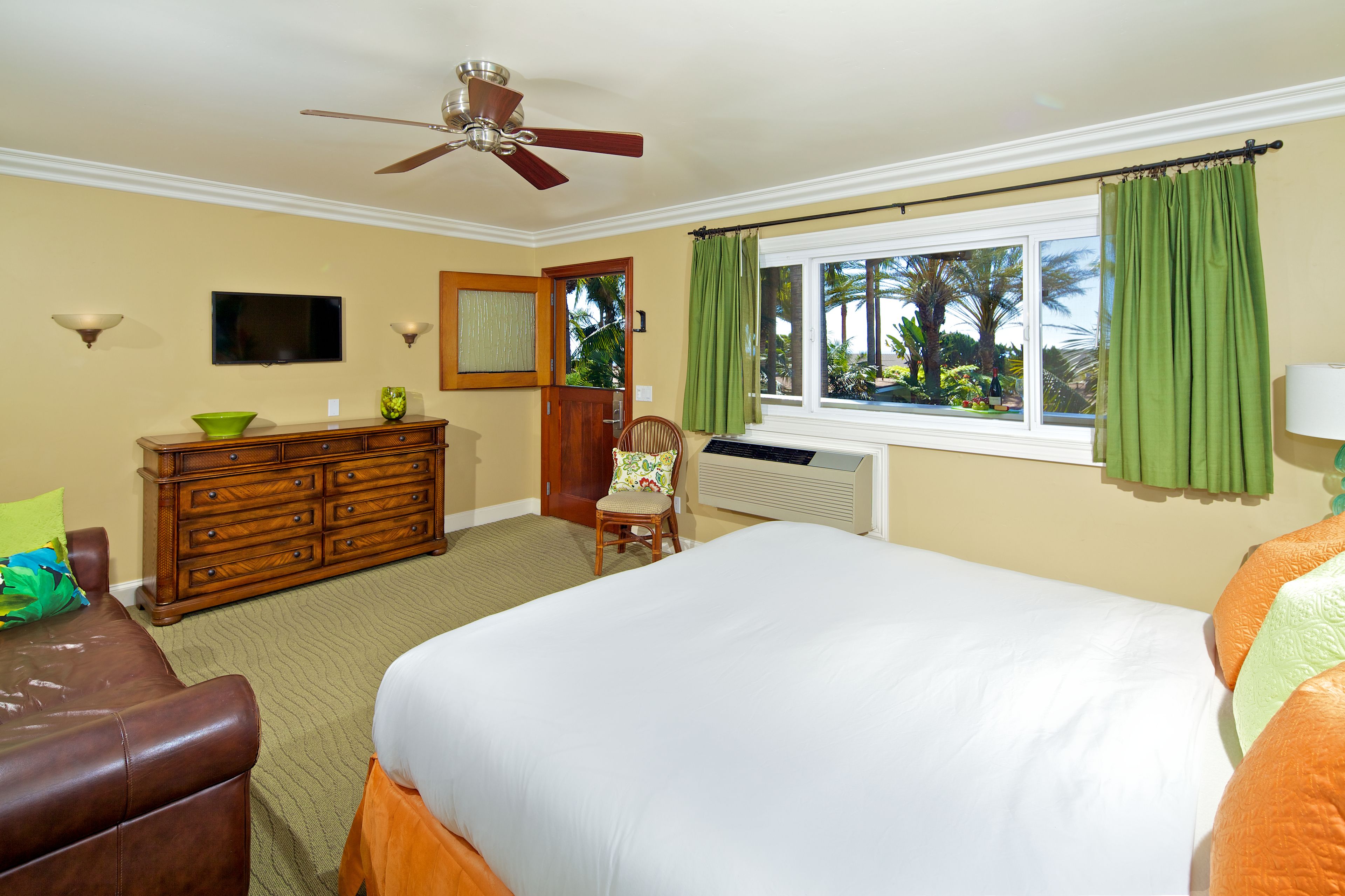 A hotel room with a bed, a dresser, a chair, a ceiling fan, a window with curtains, and a television.