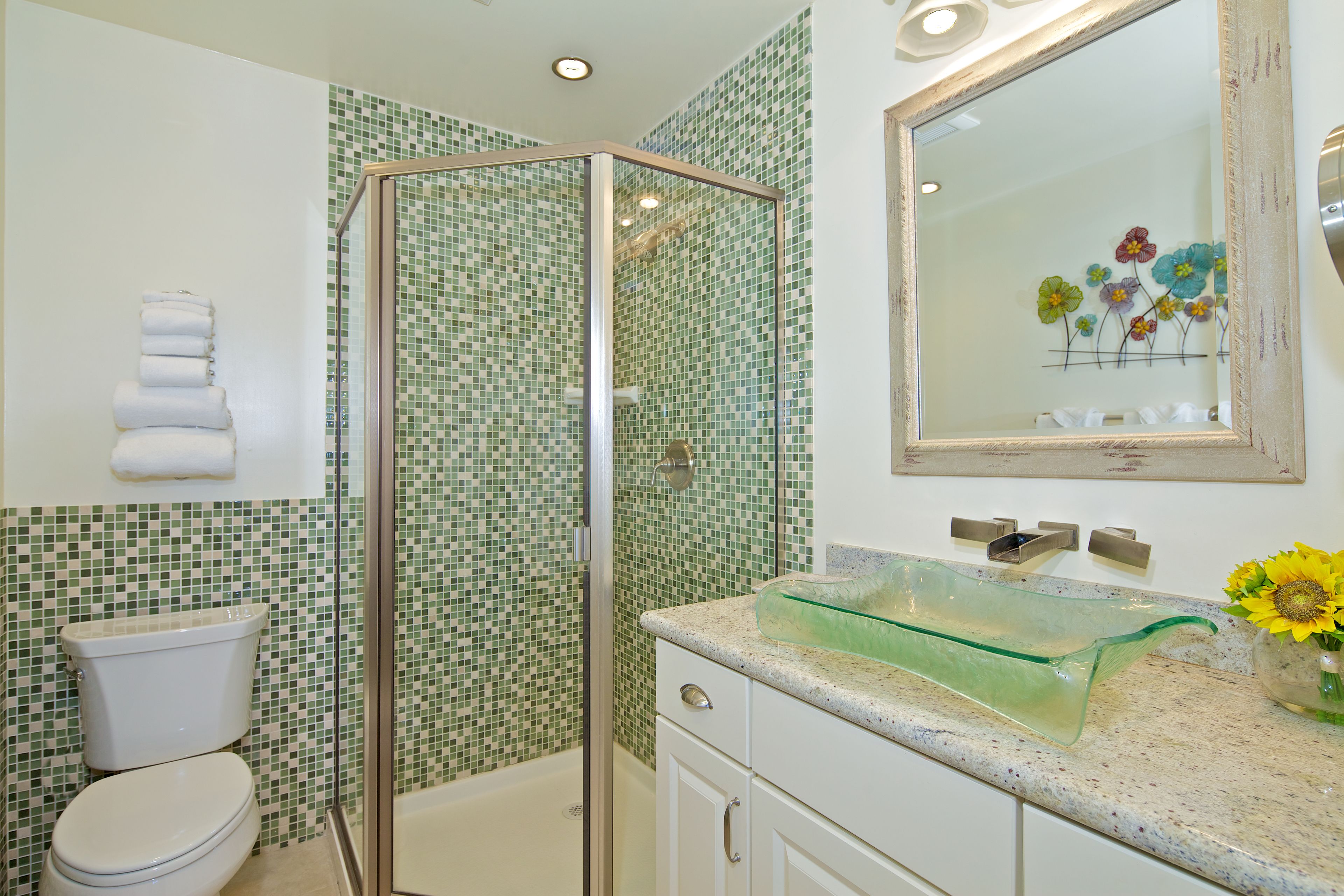 A bathroom with a glass shower enclosure, a toilet, a sink with a glass basin, and a mirror with a floral decoration.