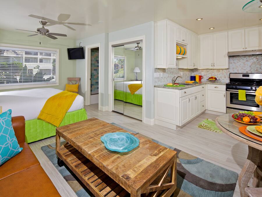 A modern hotel room with a bed, a wooden table, a kitchen area, and a balcony with a view.